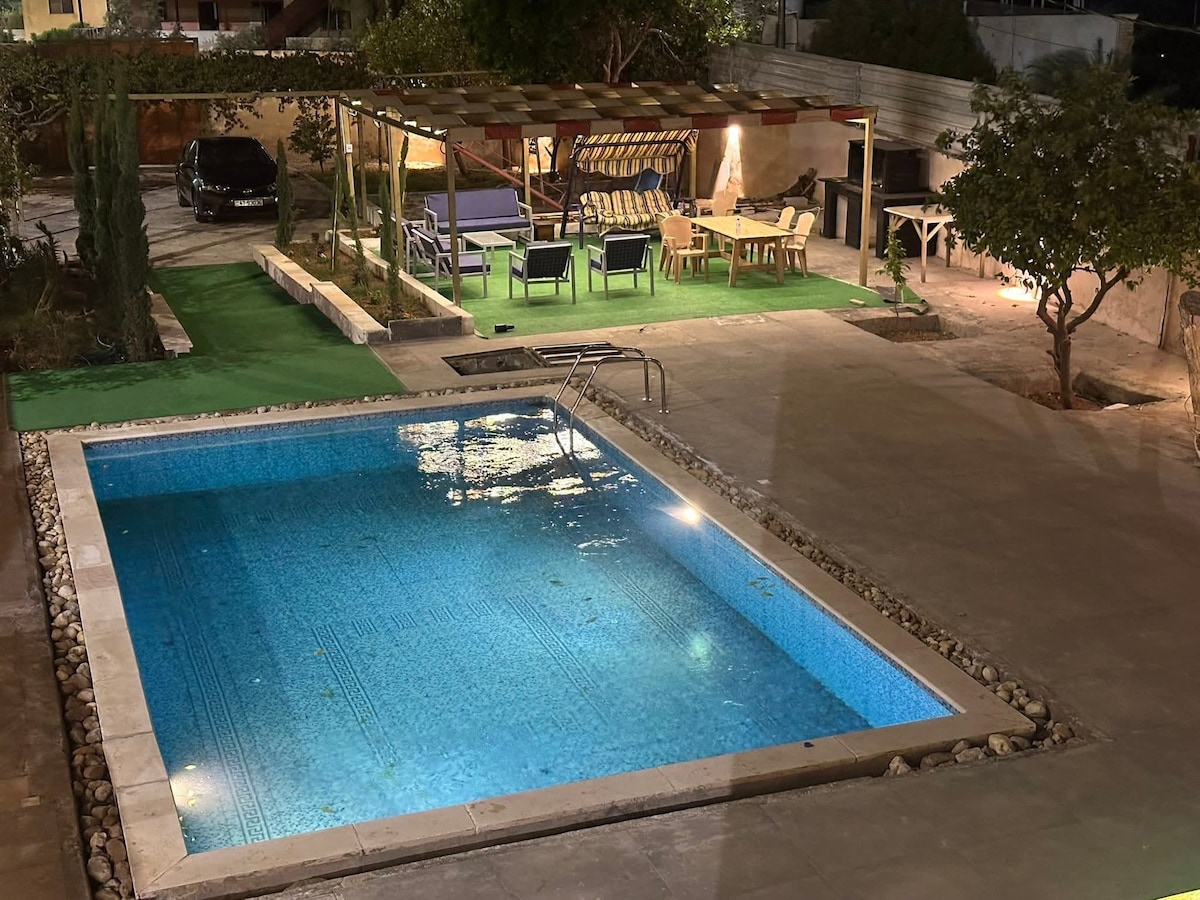 An outdoor area features a pool bordered by smooth stones, reflecting ambient light. A shaded gazebo is visible in the background, equipped with seating and lounging options. Green grass patches and trees provide a natural touch to the serene space.