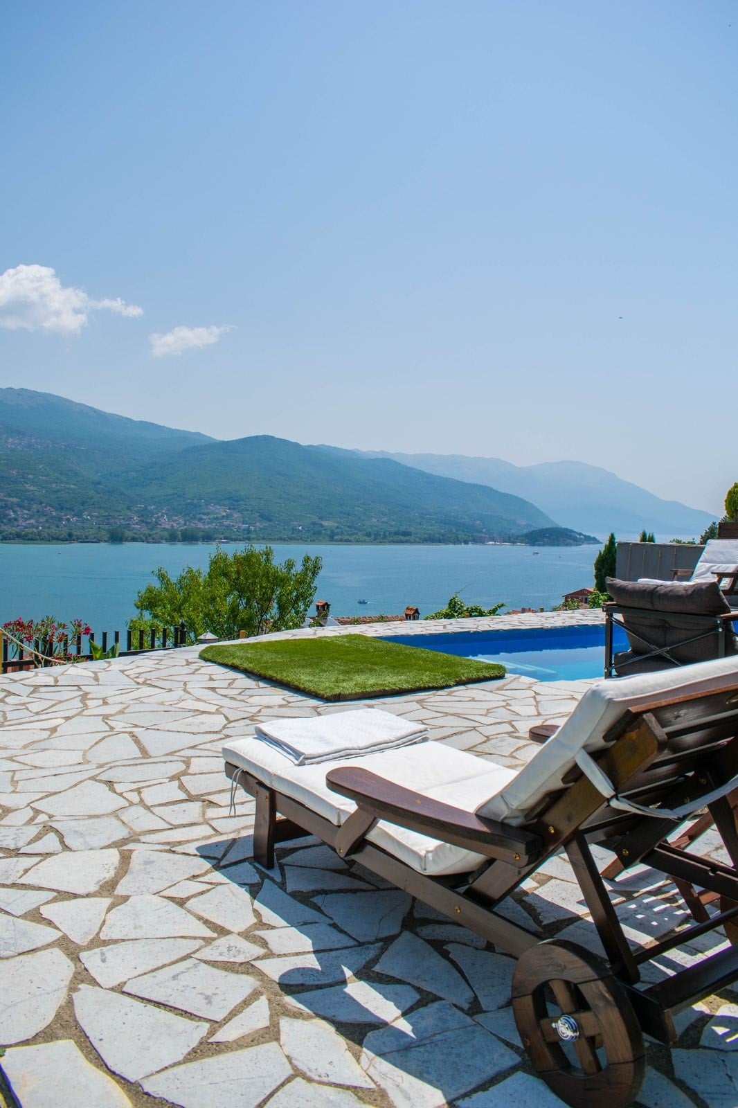 A spacious outdoor terrace showcases a stunning view of the lake and surrounding mountains. Lounge chairs are positioned beside a well-kept pool, and the patio features light-colored stone flooring. Greenery and shrubs line the edge, adding to the natural scenery.