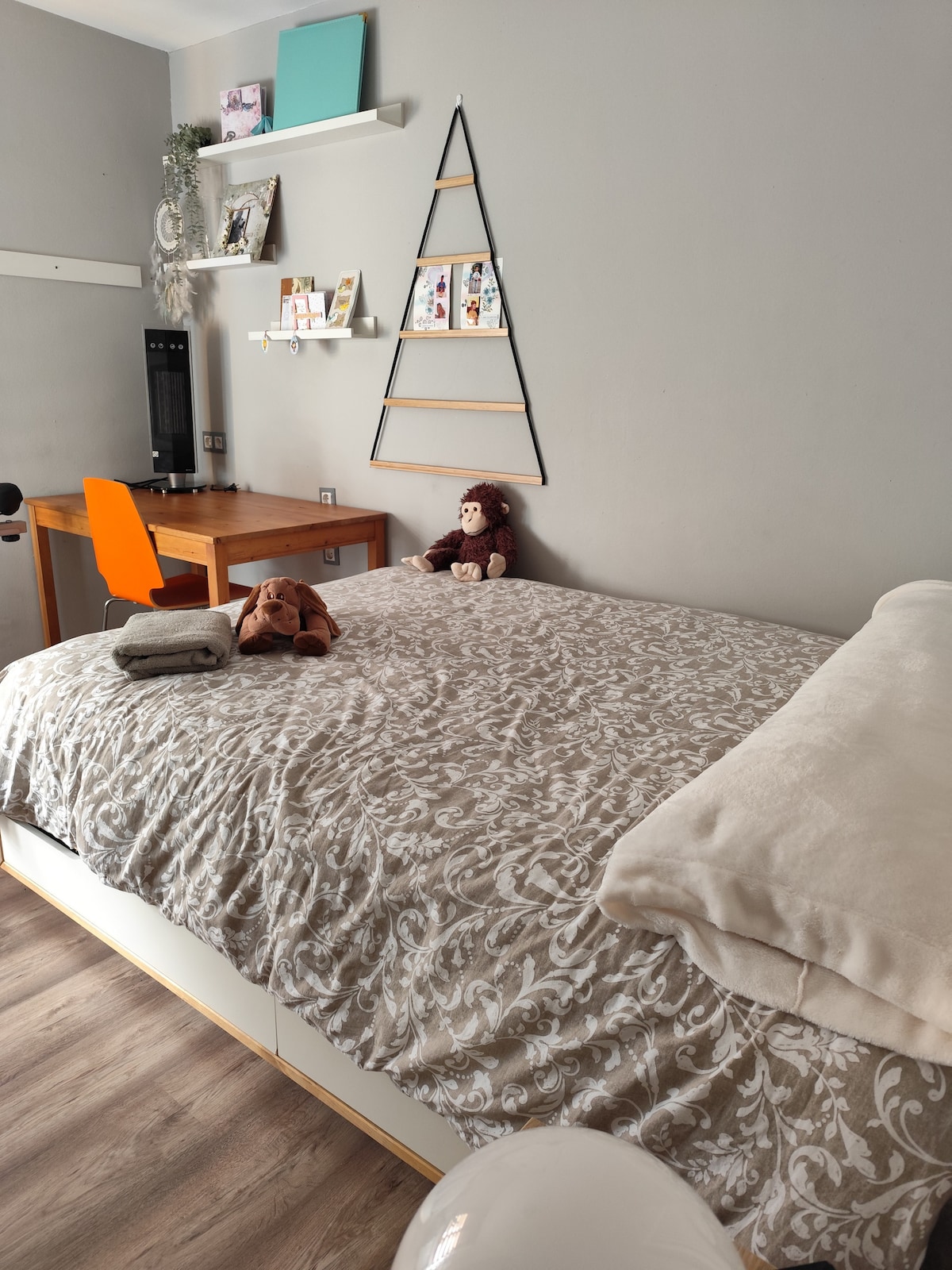 The spacious sleeping area features a large bed covered with a decorative blanket. A wooden desk is positioned nearby, with a vibrant orange chair. A plush toy sits on the bed, accompanied by neatly folded towels. Decorative shelves display personal items above the work space.