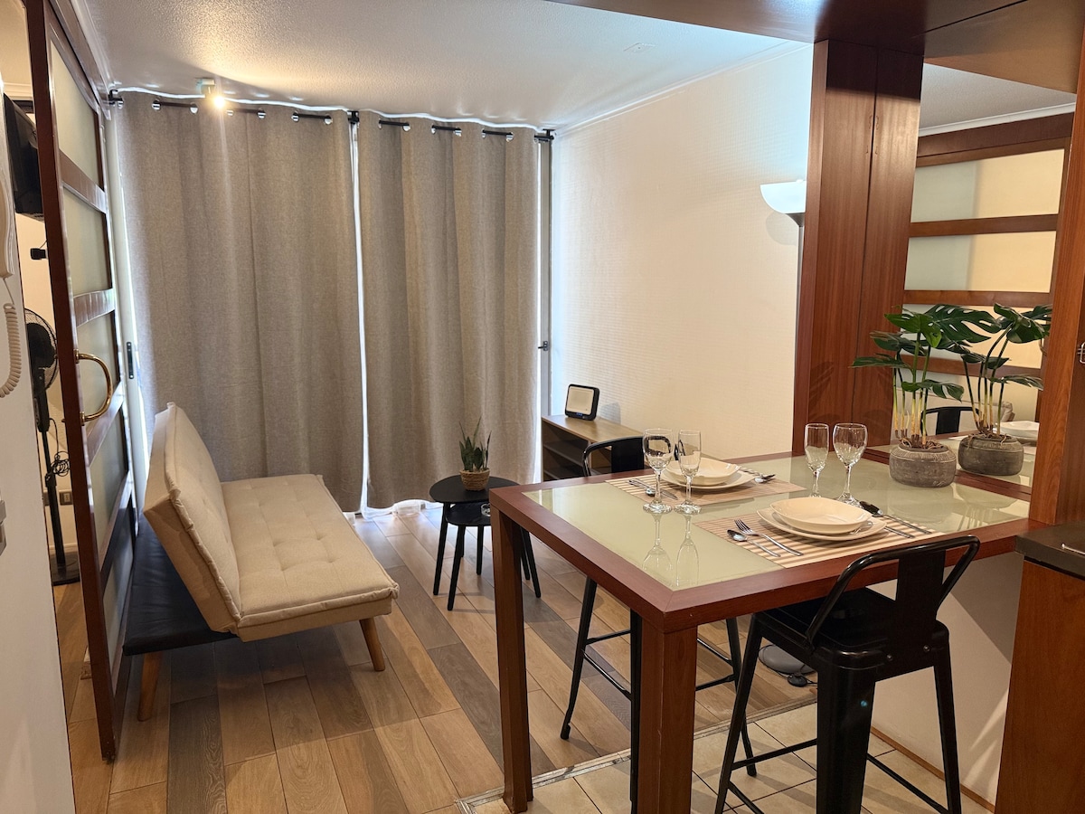 A cozy dining area features a small table set for two, with elegant dishware and glasses. A neutral-toned sofa bed is positioned nearby, complemented by a side table and a small potted plant. Natural light filters through the curtains, enhancing the space's warmth.
