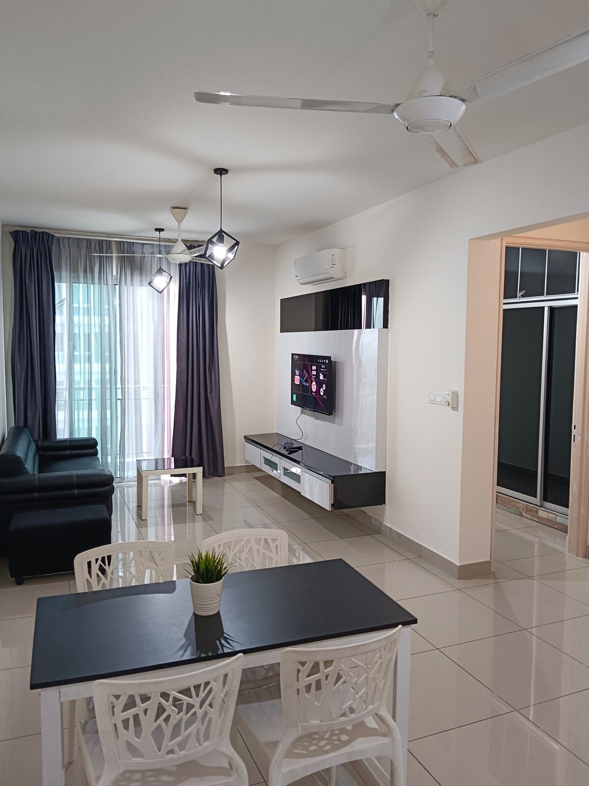A modern living area is presented with two black sofas positioned around a glass-top coffee table. A wall-mounted television is visible, while natural light enters through sheer curtains. The space is air-conditioned, featuring a ceiling fan for comfort and a decorative plant on the table.