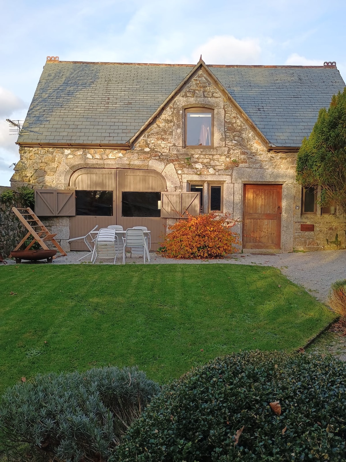 A rustic stone cottage is featured with a slate roof, surrounded by a well-maintained lawn. A private patio includes several white chairs and a wooden lounge. Colorful foliage decorates the entrance, while large windows offer ample natural light, contributing to a welcoming exterior.