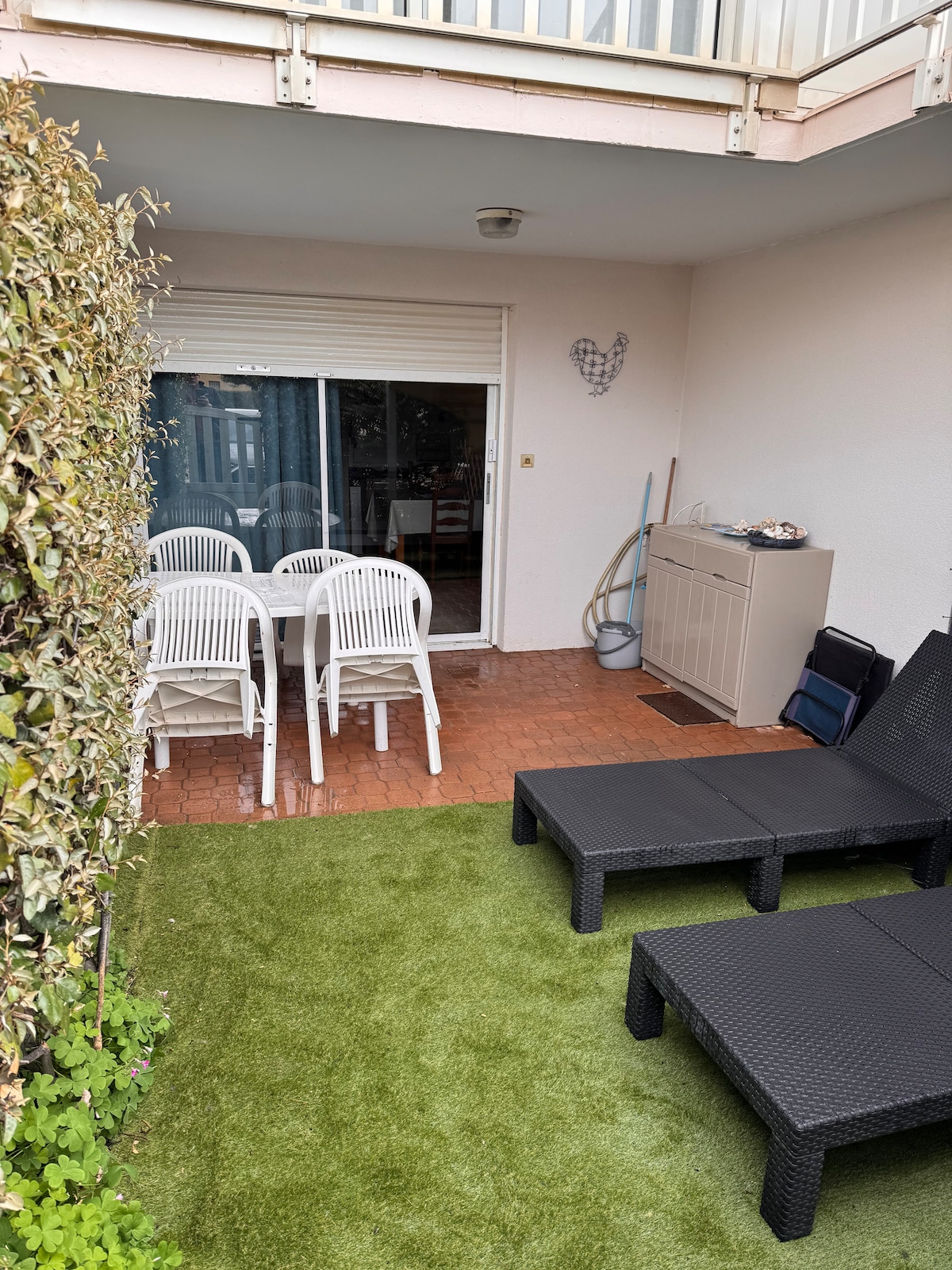 A cozy outdoor space includes a small green lawn area with two lounge chairs and a set of four white dining chairs arranged around a table. A sliding glass door leads to an interior space, while a storage console is visible nearby.