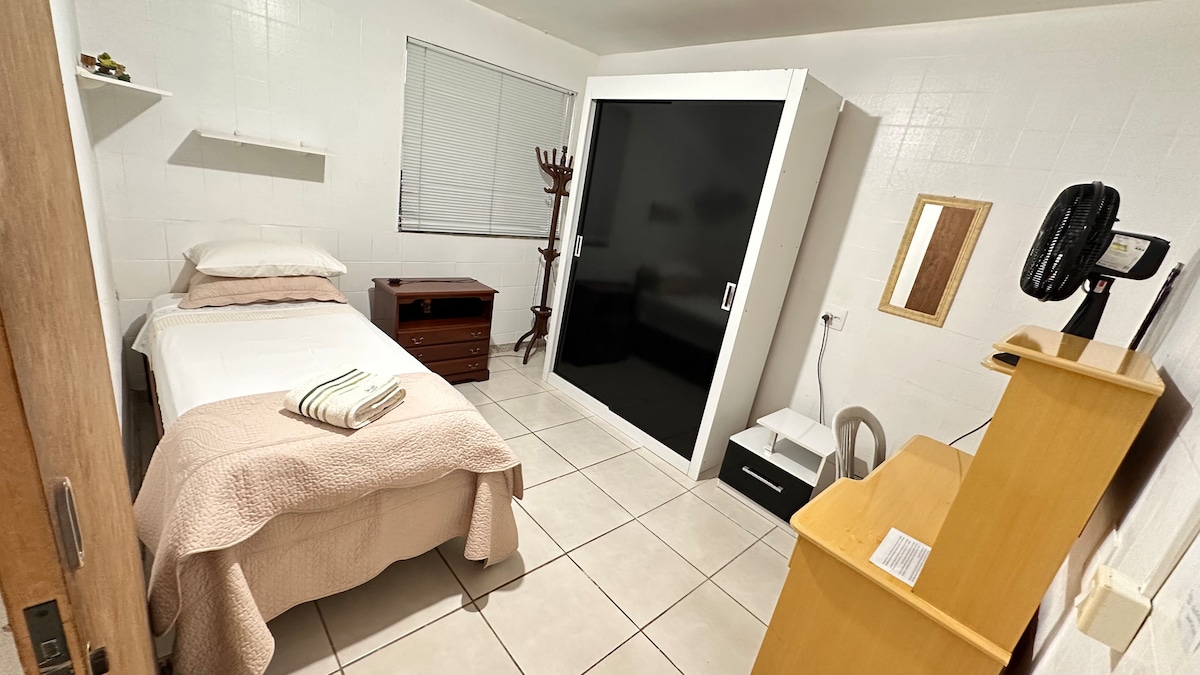 The room features a single bed with a soft cream blanket draped over it. A nightstand is placed beside the bed, and a closet with sliding doors stands against one wall. A fan and a workstation are also visible, contributing to a functional living space.