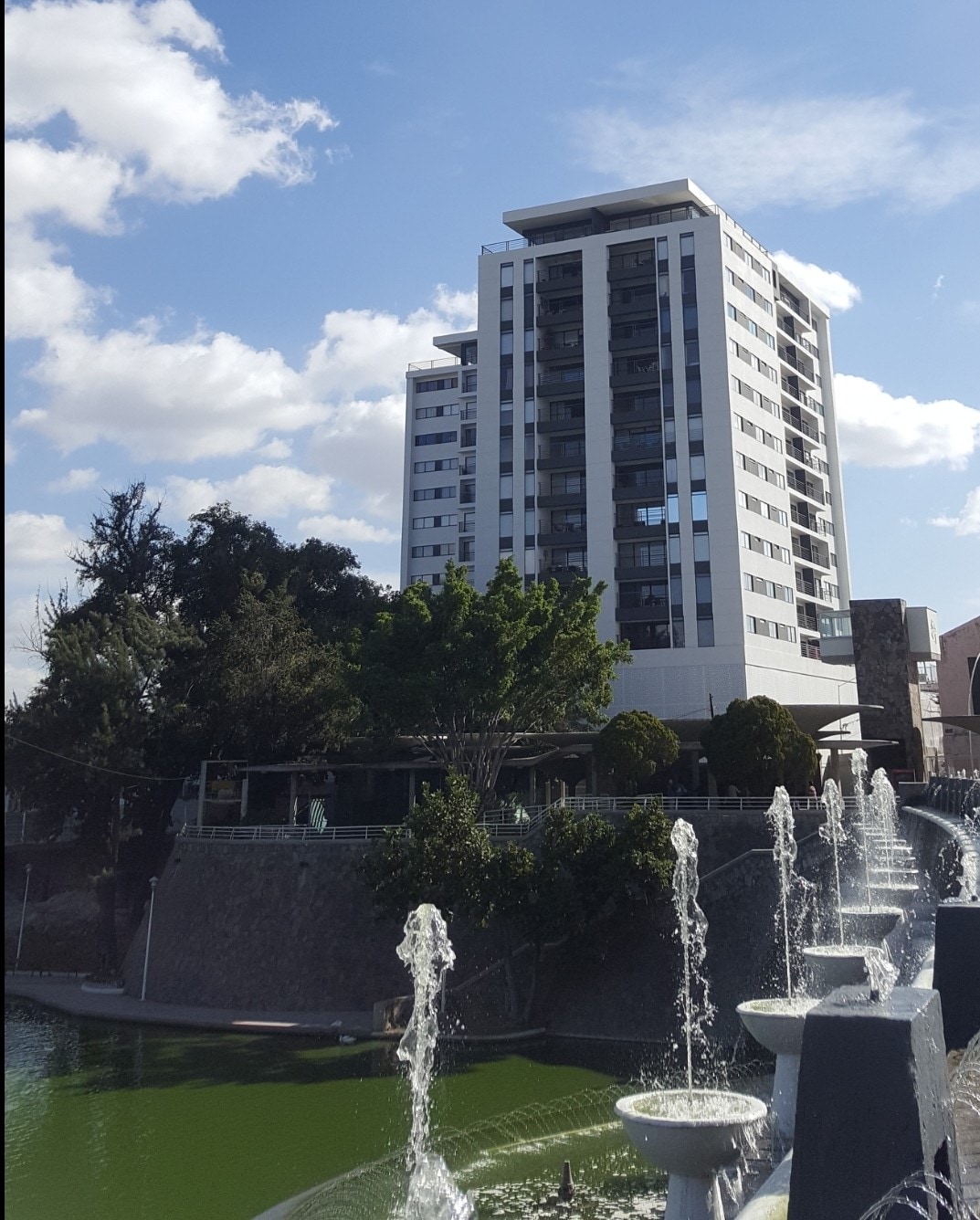 A modern multi-story building is depicted against a clear blue sky, surrounded by lush greenery. A water feature with multiple fountains is visible in the foreground, adding a serene element to the scene. The building showcases a mix of glass and white facades, reflecting light.