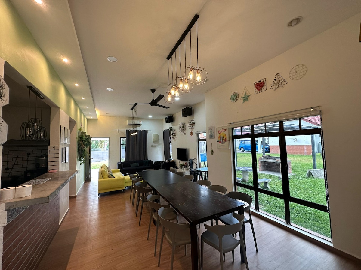 An expansive common area is featured, highlighted by a long wooden dining table surrounded by chairs. Bright lighting fixtures hang from the ceiling, and a large window offers a view of the outdoor space. A vibrant yellow sofa is positioned nearby, enhancing the lively ambiance.