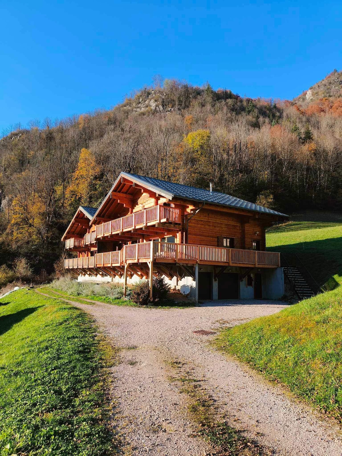 A charming chalet is positioned on a gentle slope, surrounded by lush greenery and autumn foliage. The wooden structure features multiple balconies and large windows, inviting natural light. A gravel path leads up to the entrance, bordered by well-maintained grass, providing an inviting approach.