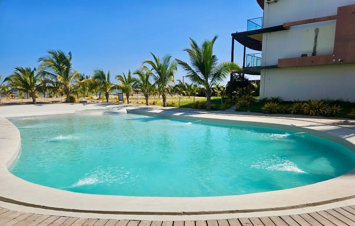 A large, circular swimming pool with inviting turquoise water is surrounded by palm trees and landscaped greenery. The pool area features a smooth deck and is positioned adjacent to a modern condominium, creating a spacious atmosphere for relaxation and enjoyment.
