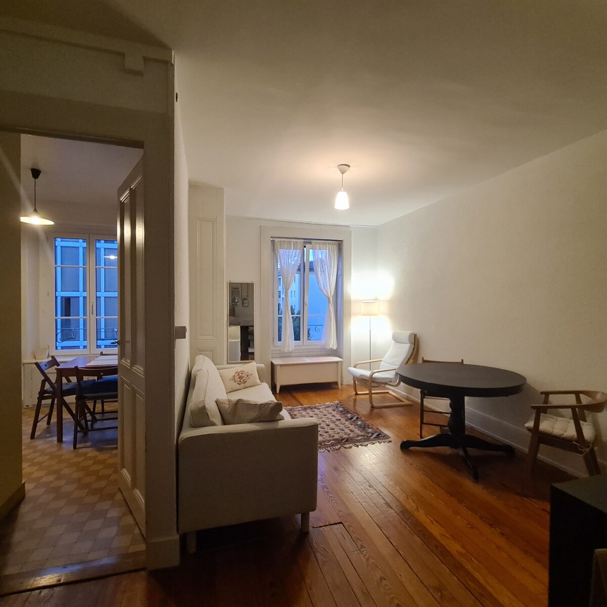 A well-lit living space features a comfortable sofa and a small round table accompanied by chairs. Natural light enters through a window covered with sheer curtains. A dining area with a larger table can be seen in the background, enhancing the room's functionality.