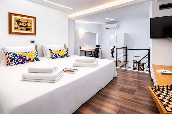 Dormitorio luminoso en el ático con cama doble, almohadas de colores, TV y vistas abiertas a la zona de estar.