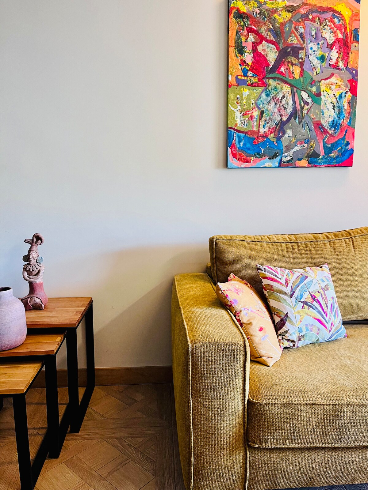 A cozy seating area is highlighted by a yellow sofa adorned with patterned cushions. Adjacent to the sofa, two wooden side tables provide a functional touch. A colorful abstract painting adds artistic flair to the wall, contributing to a lively ambience.
