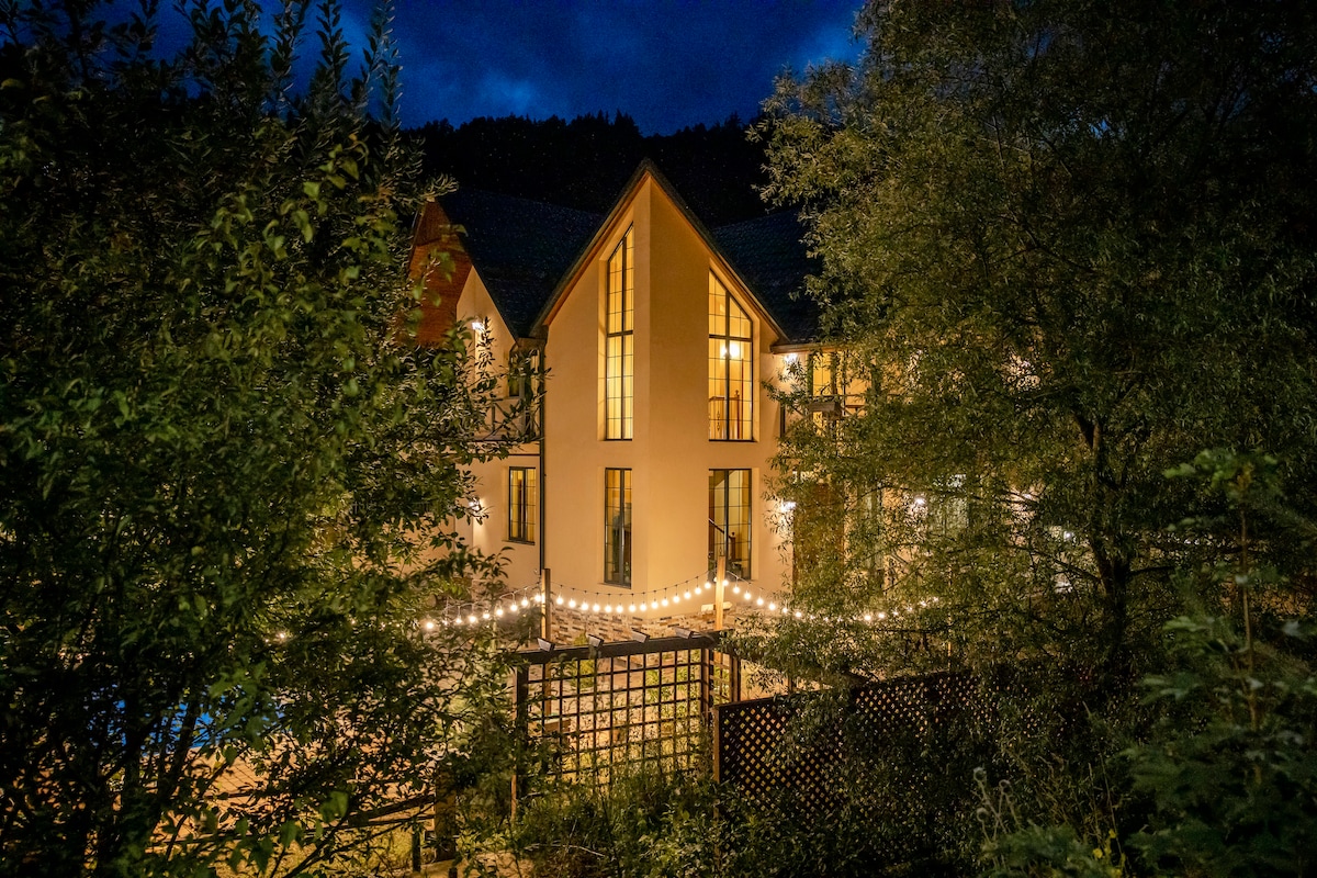 The villa is illuminated at night, framed by lush greenery. Large windows reflect the warm glow from inside, while string lights add a charming touch to the outdoor area. The forest backdrop enhances the tranquil setting, providing a connection to nature.