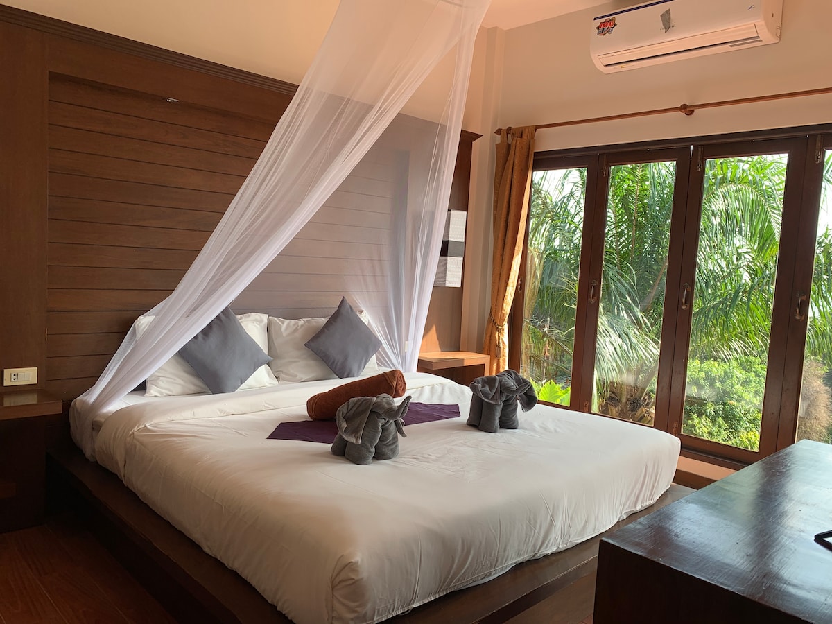 A spacious bed is draped with a white canopy, complementing the serene setting. Pillows are arranged neatly, accompanied by decorative towels shaped like animals. Large windows showcase lush greenery outside, allowing natural light to brighten the room.