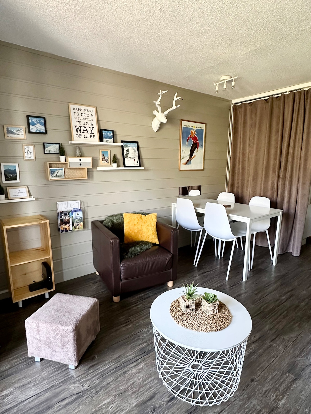 A modern living area is highlighted by a cozy armchair, complemented by a decorative cushion. The wall displays a curated selection of photos and decor. A dining table with white chairs sits adjacent, while a small ottoman and a stylish coffee table complete the space.