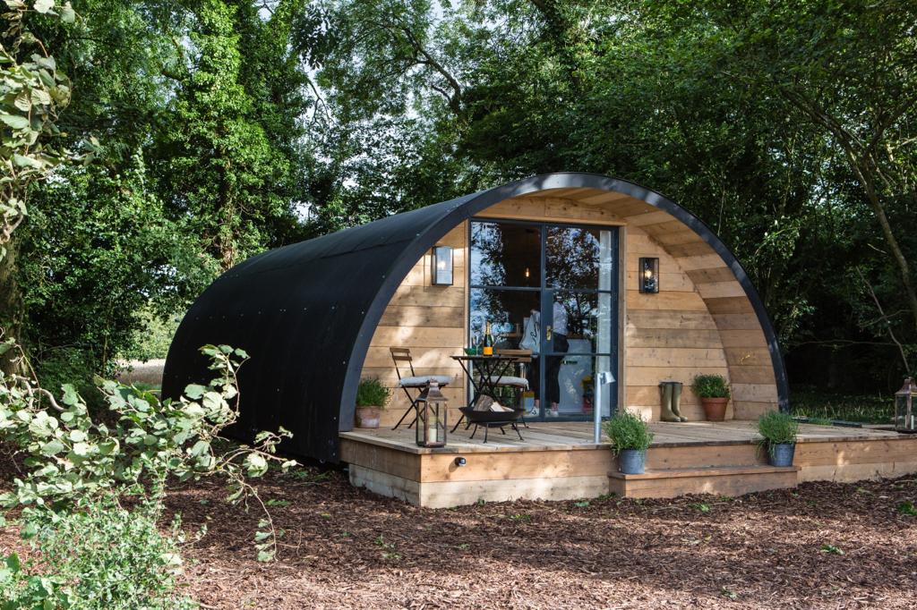 The Pigsty features a unique domed structure with a blend of dark and natural wood accents. A small outdoor seating area with a round table and two chairs is positioned at the entrance, surrounded by lush greenery and potted plants.