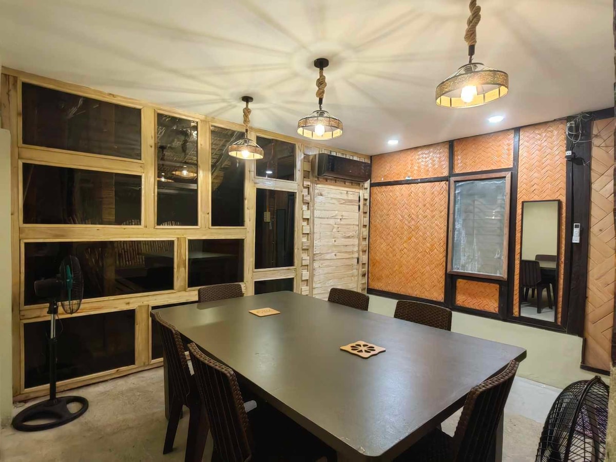A spacious dining area is highlighted by a large table surrounded by dark, woven chairs. Natural light enters through large windows, and warm pendant lights provide additional illumination. Textured walls made of bamboo add a unique touch to the inviting space.