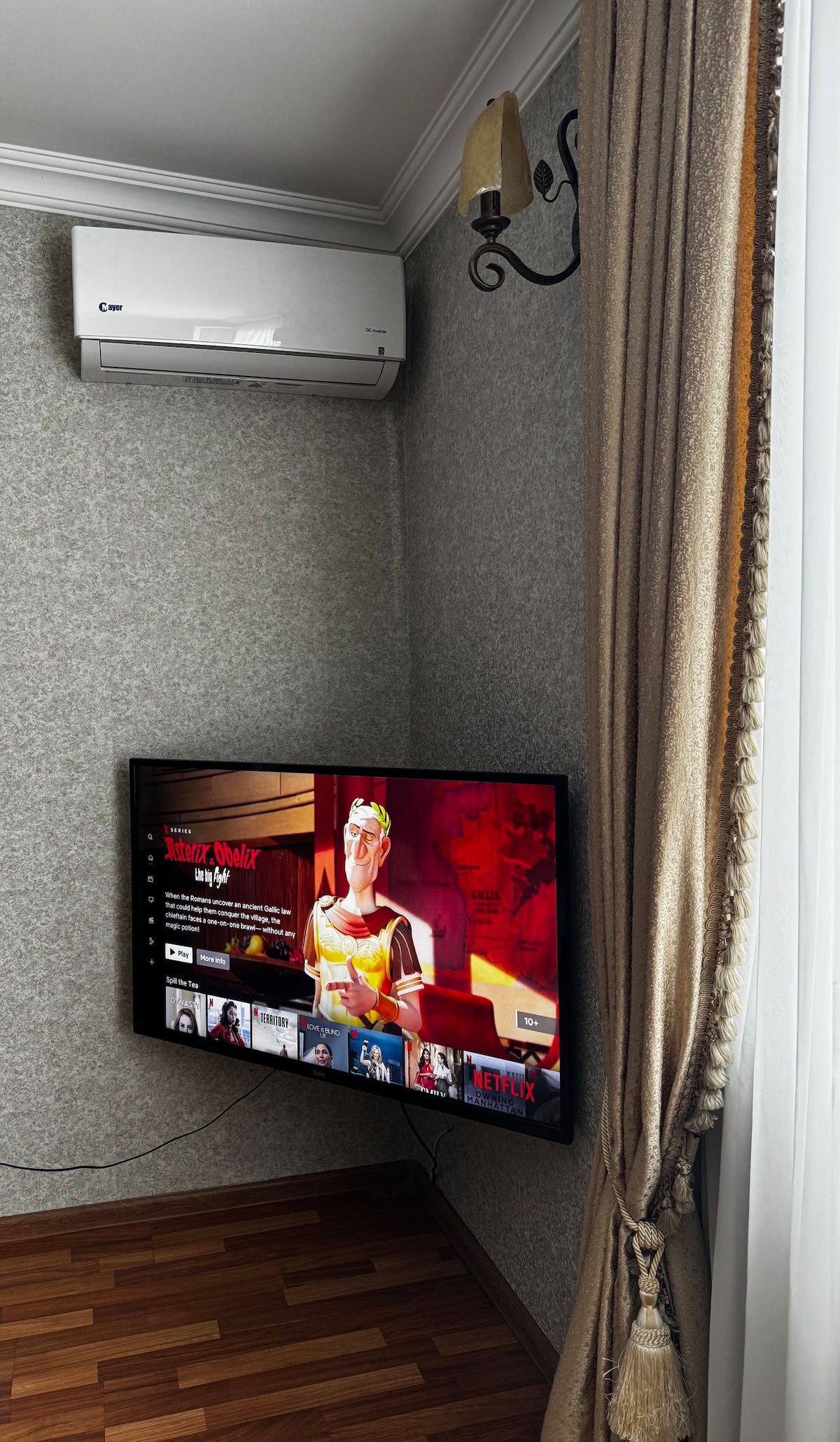 A mounted smart TV displays a streaming service interface in a cozy corner. An air conditioning unit is positioned above, while a curtain made of light fabric complements the warm-toned wooden flooring.