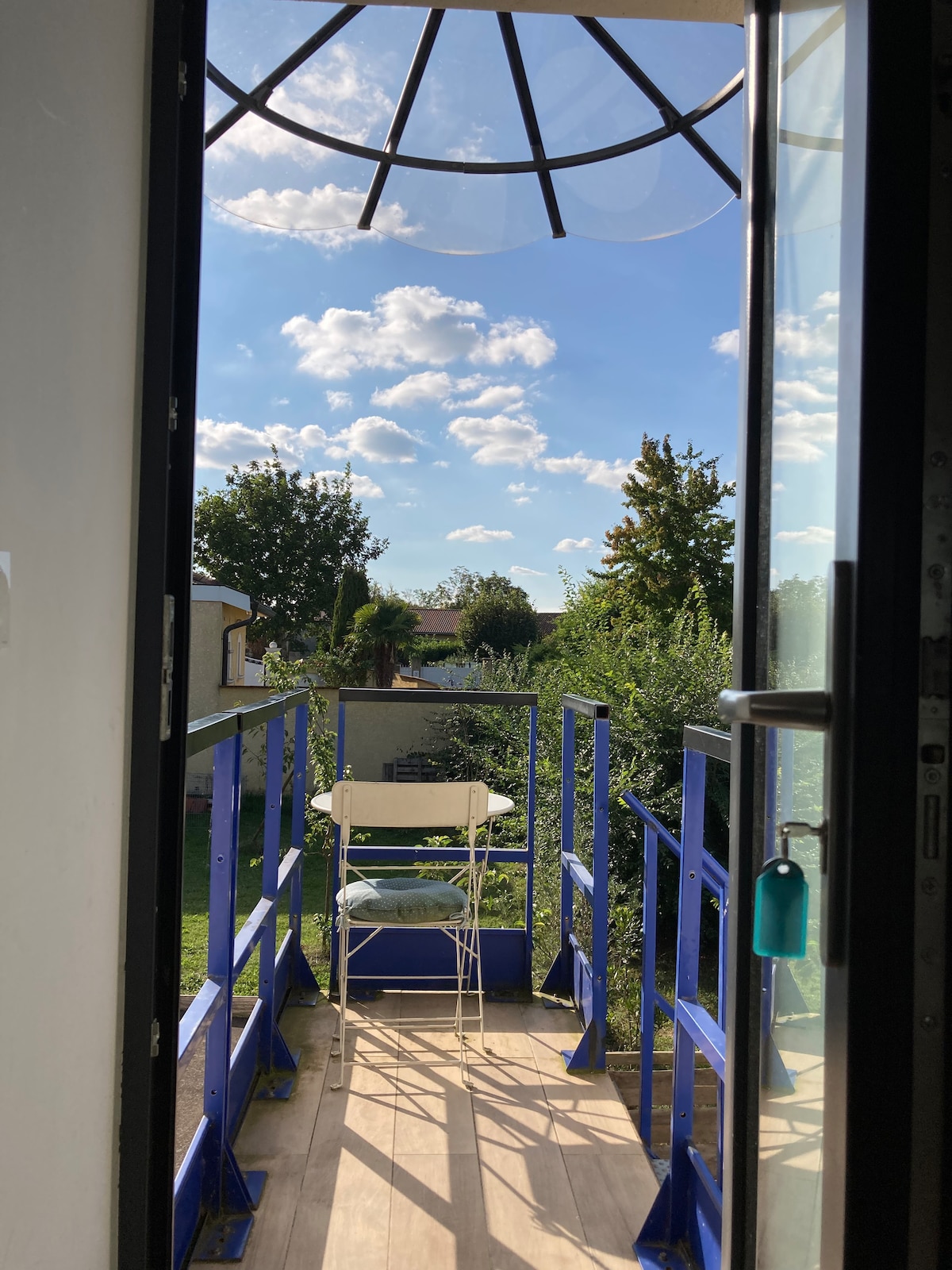 A small private terrace is visible through the open door, featuring a white table and a single chair. Lush greenery surrounds the area, and a clear blue sky with scattered clouds is seen above, contributing to the serene outdoor setting.