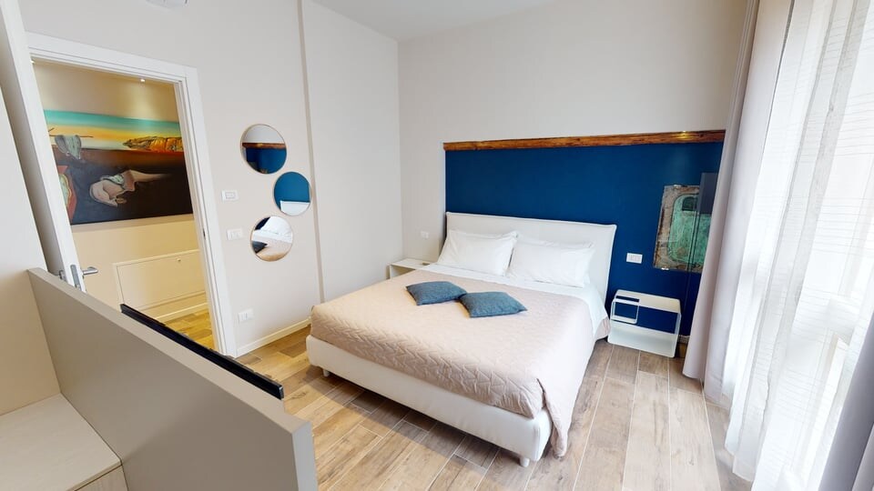 A modern bedroom features a spacious bed with light bedding. A wooden headboard is complemented by a dark blue accent wall. Natural light floods the room through large windows, enhancing the soft wooden floors. Decorative mirrors add visual interest to the space.