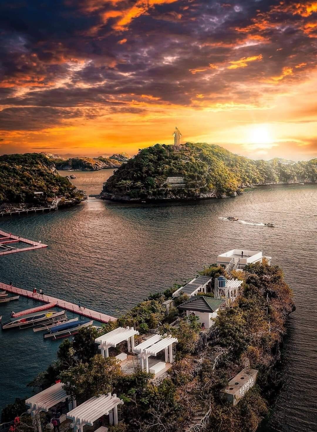 A scenic view of the Hundred Islands features lush greenery and rocky formations rising from sparkling waters. A vibrant sunset casts warm hues over the landscape, with a statue visible atop a distant hill. Boat docks and small vessels are situated near the shoreline.
