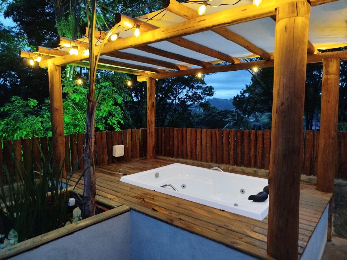 An outdoor space features a wooden pergola illuminated by soft string lights. A private hot tub is situated on a wooden deck, surrounded by lush greenery and a wooden fence, creating a serene atmosphere for relaxation.