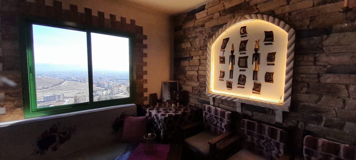 A cozy interior space features a large window with a view of the Beqaa Valley. Stone wall accents add texture, while a display niche holds various framed items. Comfortable seating is arranged to enjoy the scenery, with soft textiles enhancing the setting.