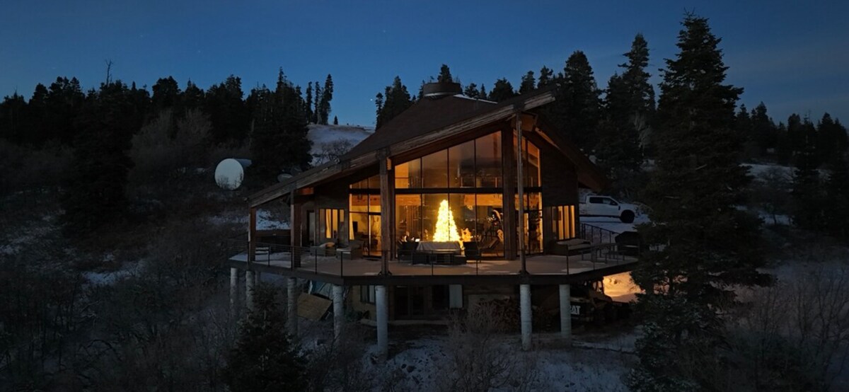 The home is positioned on elevated land, showcasing large glass panels that reflect the warm glow from inside. Surrounded by trees and snow, the architectural design harmonizes with the natural setting, creating a cozy ambiance amidst a serene landscape.