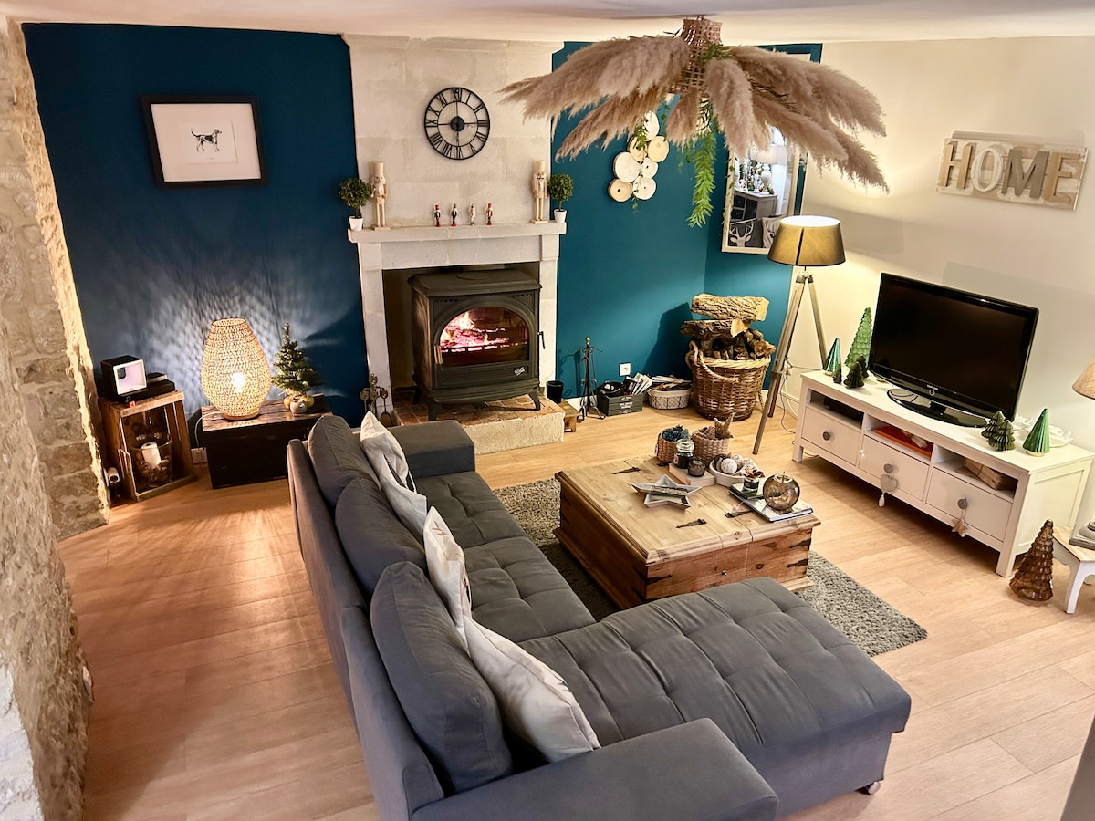 A living room features a cozy gray sofa positioned around a rustic wooden coffee table. A decorative lamp and a television are visible, with a fireplace providing warmth. Earthy tones in the decor complement a stone wall, creating a welcoming atmosphere.