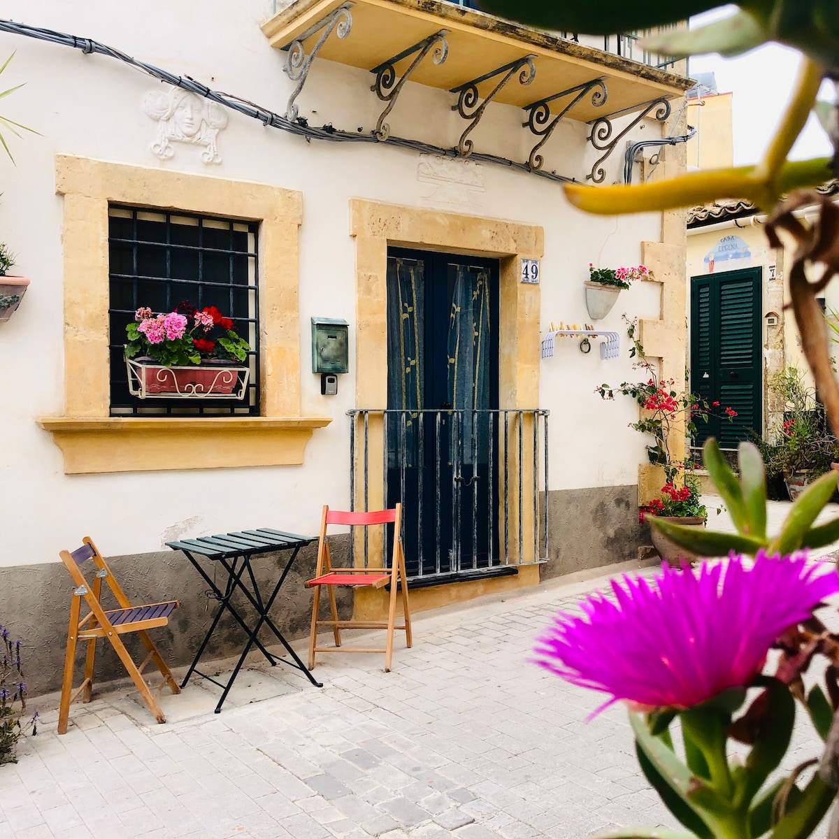 A charming exterior view features a yellow building with decorative elements. A blue door is framed by a stylish wrought-iron gate. Potted flowers adorn the window, while a small table and two chairs are placed on the sidewalk, inviting relaxation.