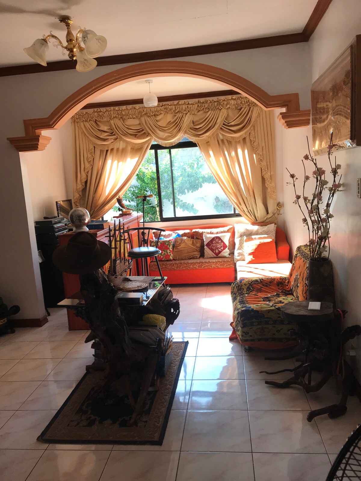 A cozy living area is highlighted by a large window draped with elegant curtains, allowing natural light to fill the space. An orange sofa and various decorative cushions create a welcoming atmosphere. A small table and plants add to the room's charm.