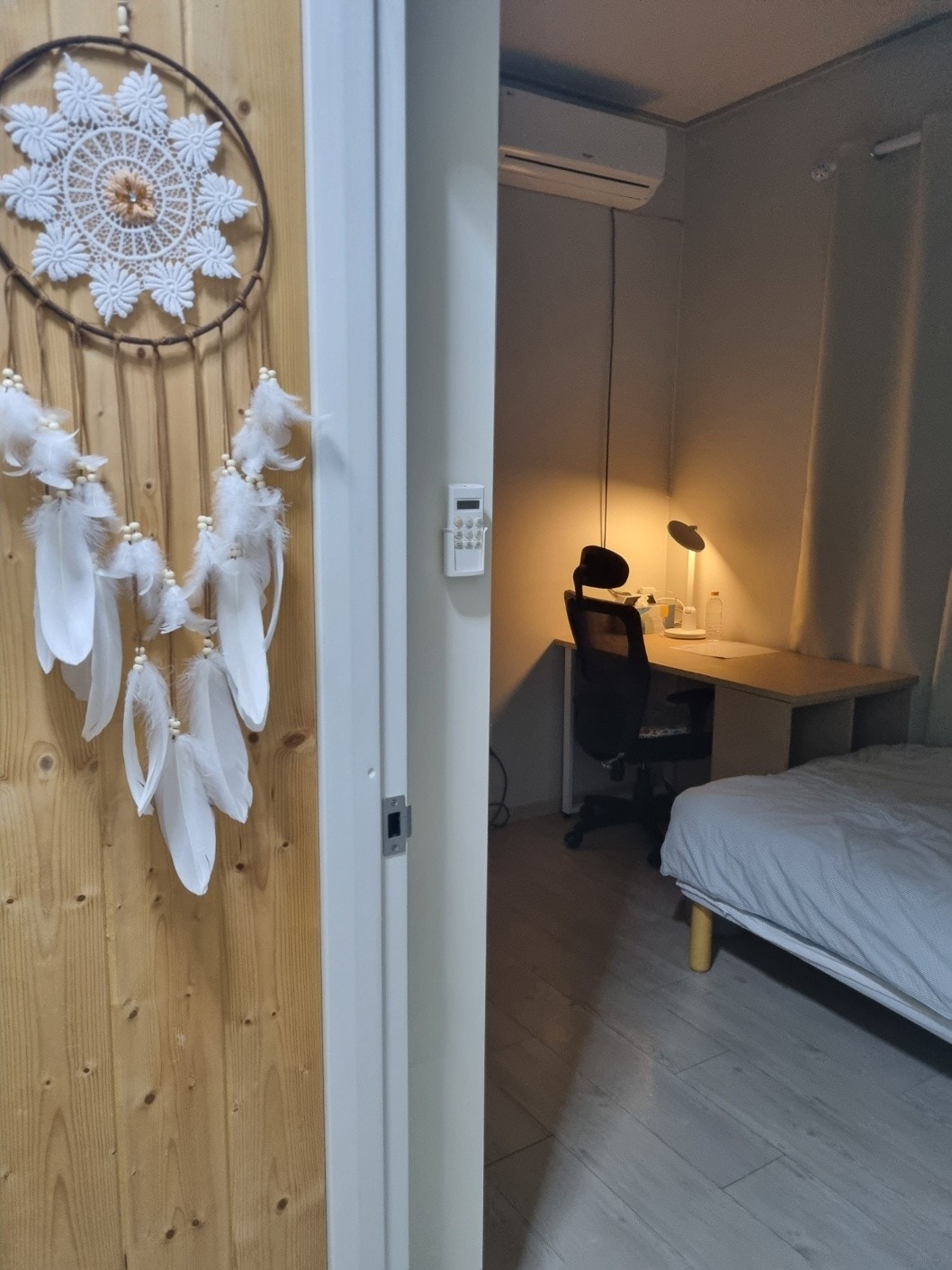 A soft-lit space features a cozy sleeping area with a bed and a minimalist desk setup. A wooden door with decorative dreamcatchers adds texture, while curtains frame the window, allowing for warm light to enhance the room's ambiance.