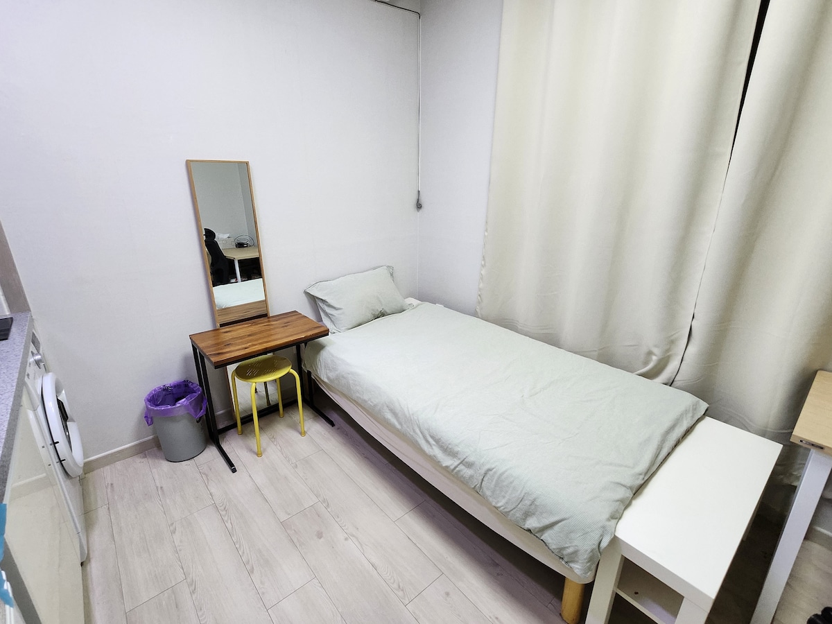 A simple sleeping area is presented, featuring a spacious bed with light bedding against a neutral wall. A small table with a yellow chair and a full-length mirror is positioned nearby. Natural light filters through a partially closed curtain, enhancing the room's brightness.