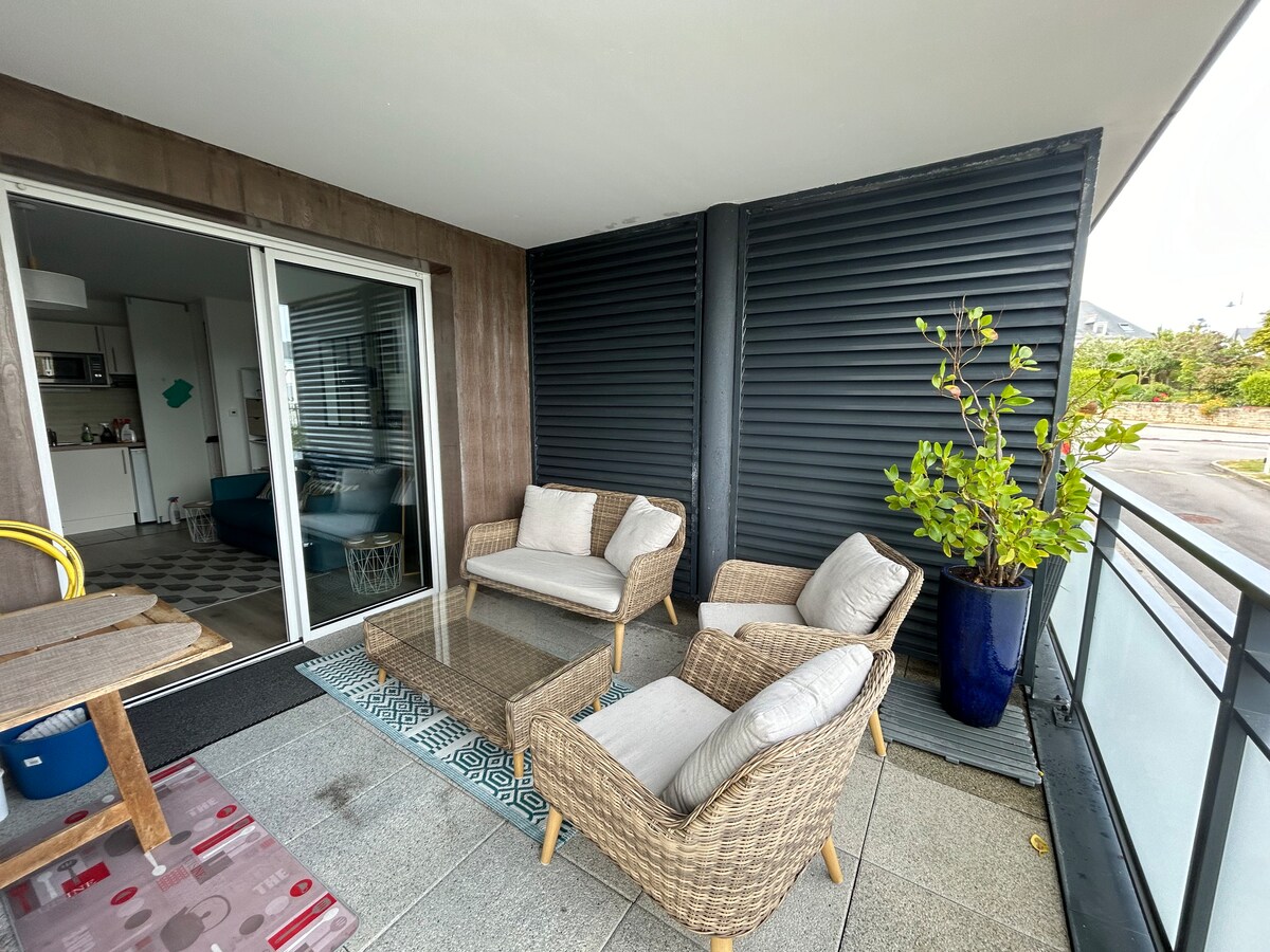 A terrace area is featured, furnished with two comfortable armchairs and a glass-topped table. A potted plant adds a touch of greenery. Large glass doors provide access to the interior space, offering a seamless transition between indoor and outdoor areas.