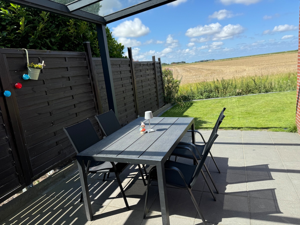 A shaded outdoor terrace features a rectangular table surrounded by four chairs. The scene is framed by wooden fencing and a well-maintained lawn. In the distance, golden fields stretch under a bright blue sky, dotted with soft clouds.
