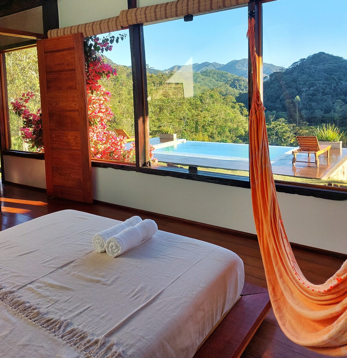 A serene bedroom features large windows offering a view of lush mountains. A spacious bed with fresh towels is complemented by a vibrant hammock nearby. Outside, a tranquil infinity pool and sun-lounger can be seen, enhancing the connection to nature.