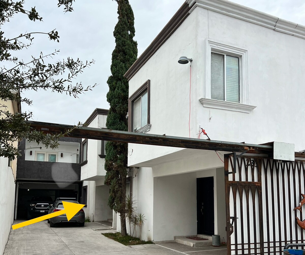 An exterior view of a two-story residence is shown, featuring a white facade and a covered parking area. A tall, green tree is visible next to the structure, and a secured gate is partially open, providing access to the driveway.