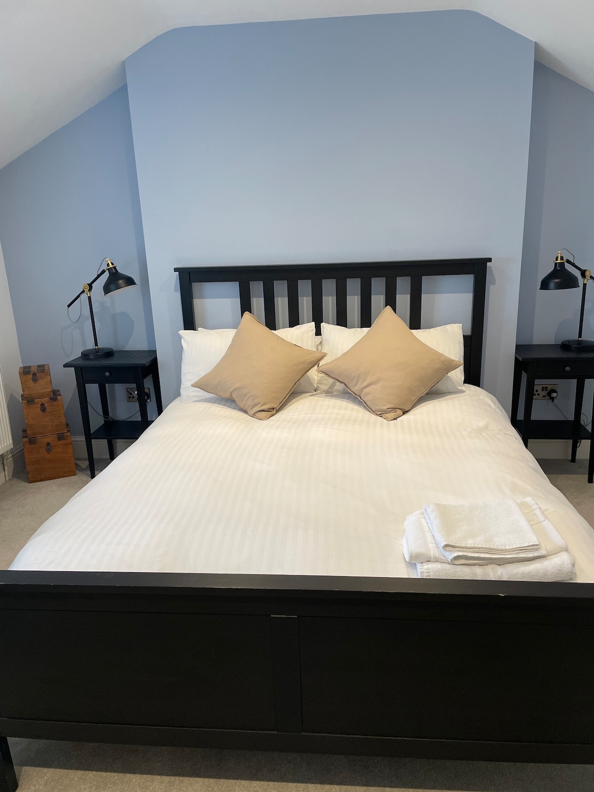 The sleeping area features a spacious bed with crisp white linens and two decorative pillows. Black bedside tables with reading lamps flank the bed, and a folded towel is neatly placed at the foot of the bed, while a light blue wall creates a calming backdrop.