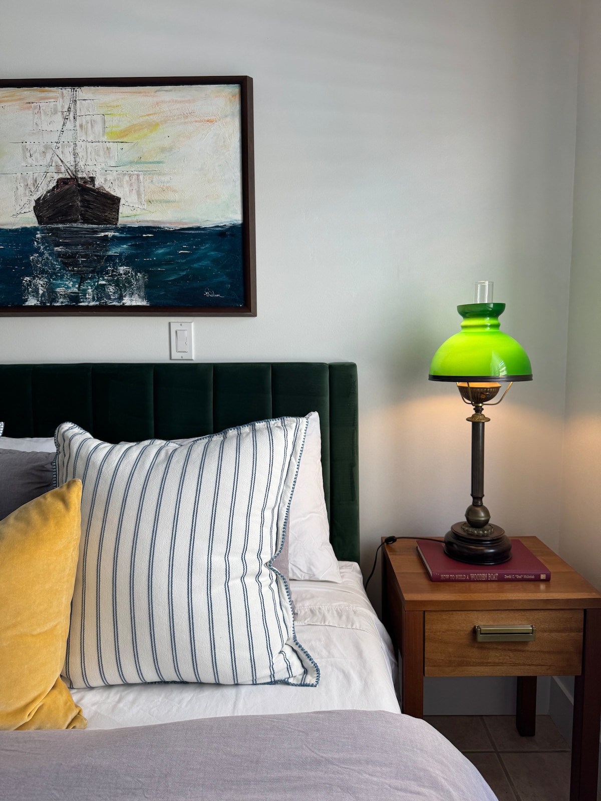 A bed with a plush green headboard is accompanied by decorative pillows in striped and solid colors. A bedside table with a wooden finish supports an antique-style lamp featuring a green shade. Artwork depicting a ship in the ocean is hung above the bed.
