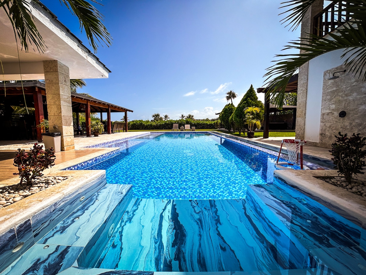 A large pool features a mosaic tile design and is surrounded by tropical foliage. Two lounge chairs are visible at the shallow end, while a cascading water feature enhances the serene atmosphere. The pool area is complemented by a spacious patio and glimpses of the landscaped gardens.