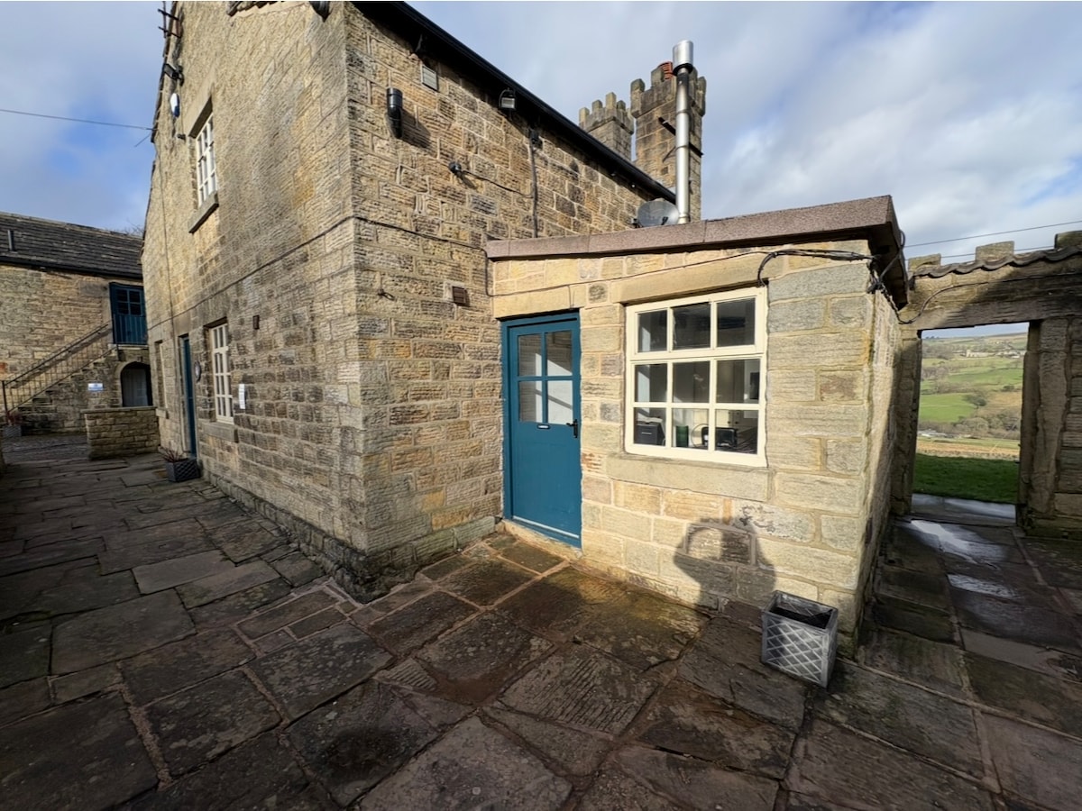 The exterior of a stone building showcases a blue door and several windows, framed by rustic stone walls. A pathway with flagstone tiles leads to the entrance, while views of the countryside can be glimpsed in the background under a partly cloudy sky.