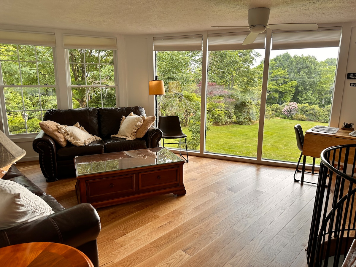A bright living room features a leather sofa and armchair arranged around a wooden coffee table. Large glass sliding doors allow natural light to fill the space, offering views of the lush greenery outside. A ceiling fan hangs above for added comfort.