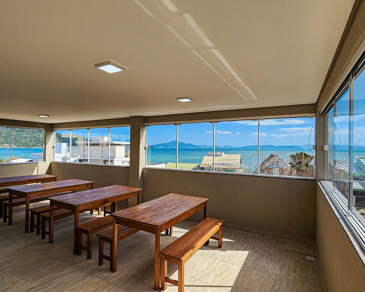 An open dining area is presented with multiple wooden tables and benches, positioned near large windows offering a panoramic view of the sea and surrounding landscape. Natural light enhances the space, creating a bright and welcoming environment.