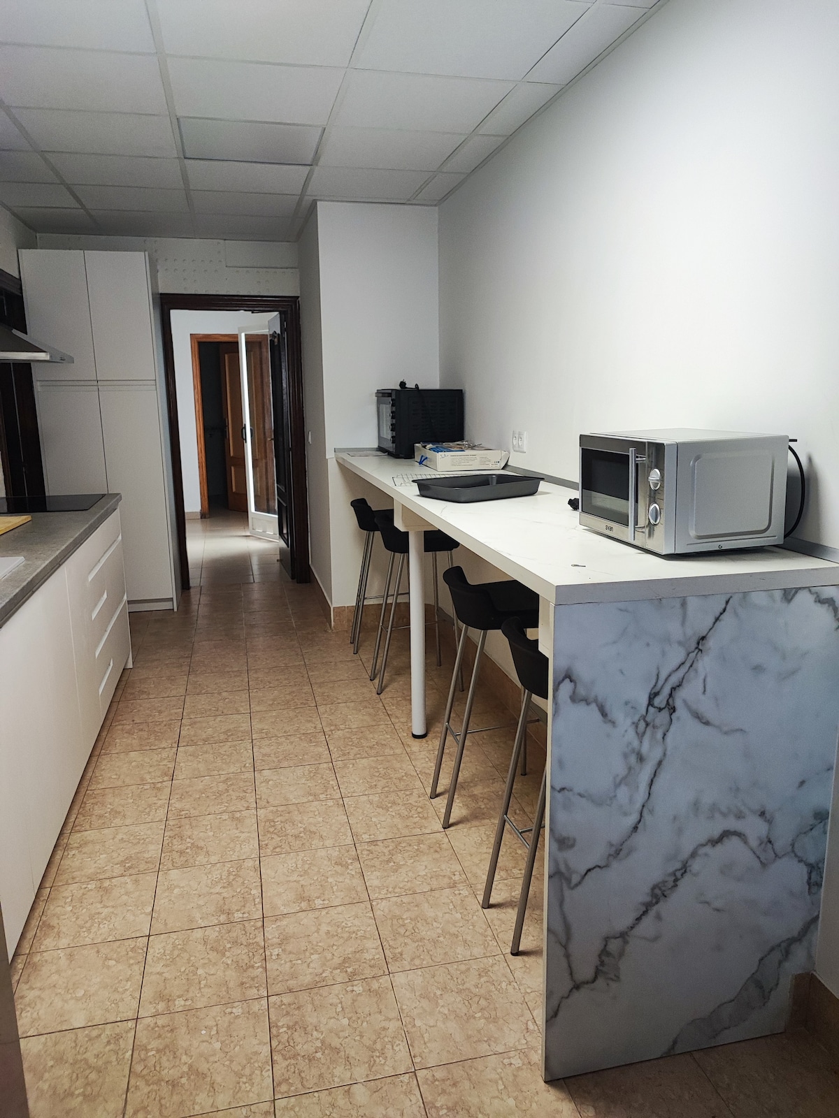 A narrow kitchen area is equipped with modern cabinetry in a light finish. High stools are positioned at a sleek, marble-topped counter. Surfaces are kept clear, with a microwave visible on the counter. A door leads to another room in the background.