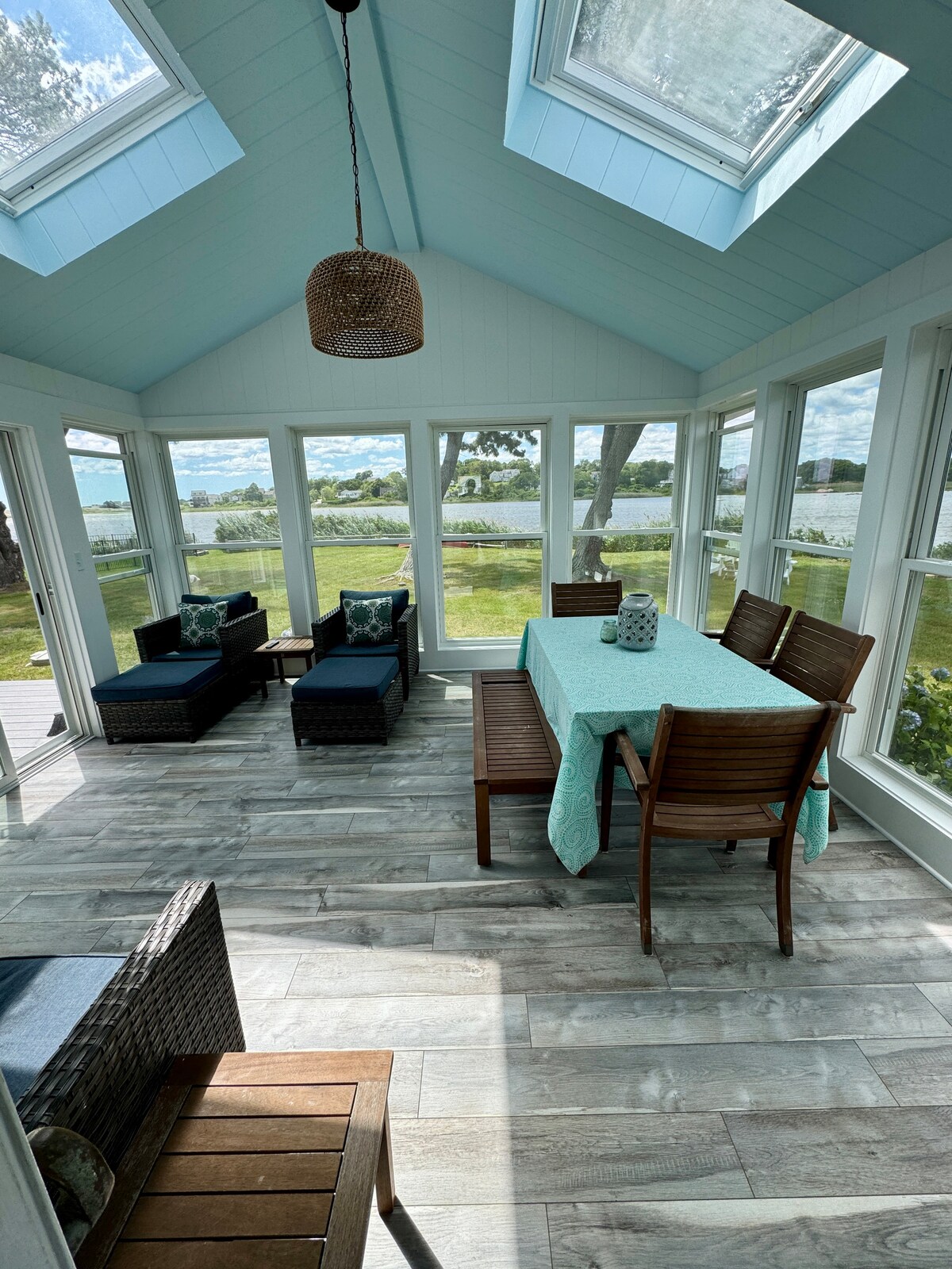 A sunroom with large windows offers views of the pond and surrounding landscape. A wooden dining table is set with a light blue tablecloth, complemented by several wooden chairs. Two cushioned lounge chairs are positioned nearby, enhancing the serene ambiance of the space.