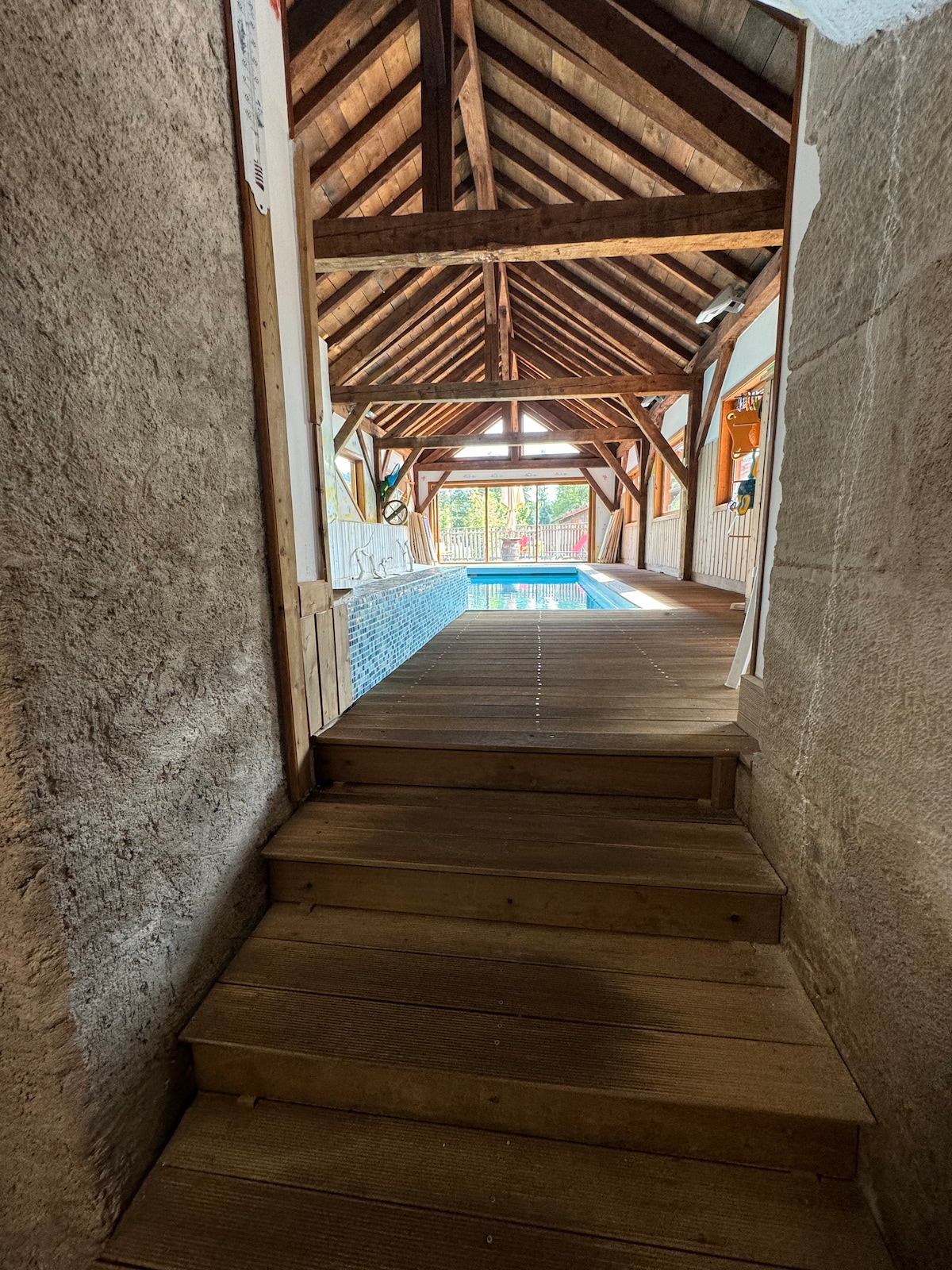 A wooden staircase leads up to a spacious indoor pool area, featuring high wooden beams and large windows. Natural light floods the space, highlighting the tranquil blue water of the pool. The surroundings blend rustic charm with modern comfort.