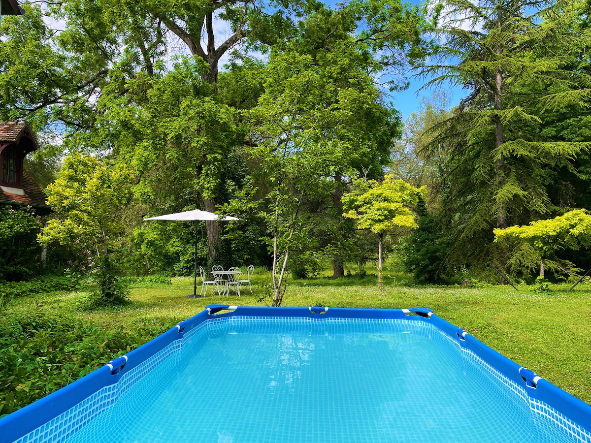 A swimming pool with clear blue water is framed by lush greenery and tall trees. A small table and chairs are positioned under a white umbrella nearby, providing a shaded spot to relax in the garden.