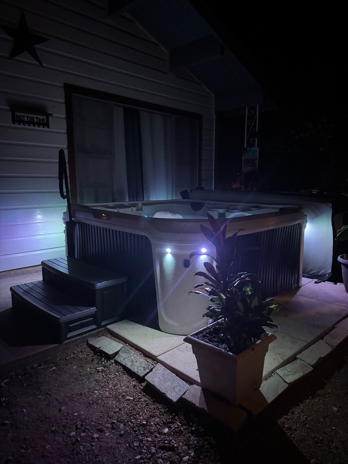 A hot tub is situated on a stone patio illuminated with soft lighting, casting a serene glow. Steps lead up to the tub, which is surrounded by potted plants. The nearby window adds a subtle reflection to the scene, enhancing the nighttime ambiance.
