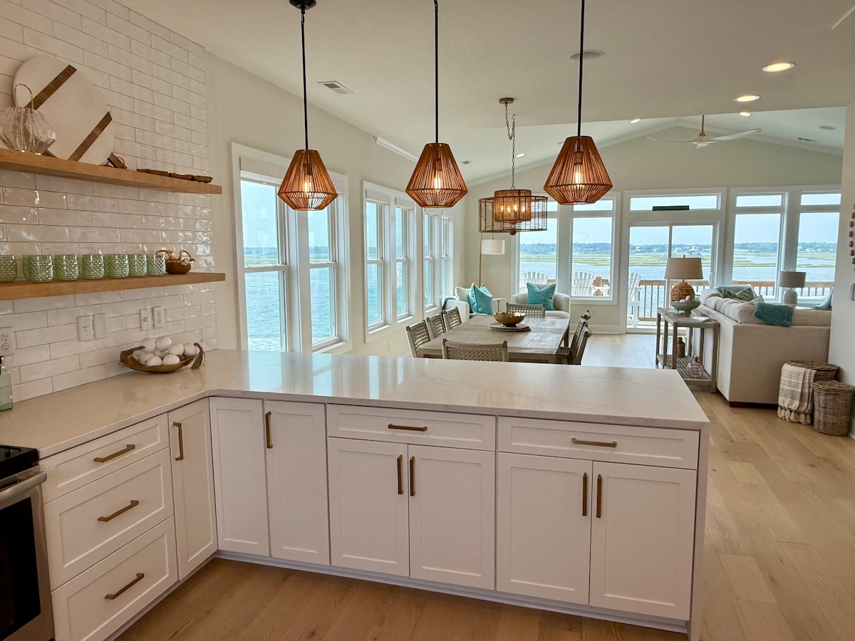 The open-concept kitchen features white cabinetry and light countertops, complemented by woven pendant lights. Large windows provide panoramic views of the water, while a seating area with a dining table and comfortable couches can be seen in the background.
