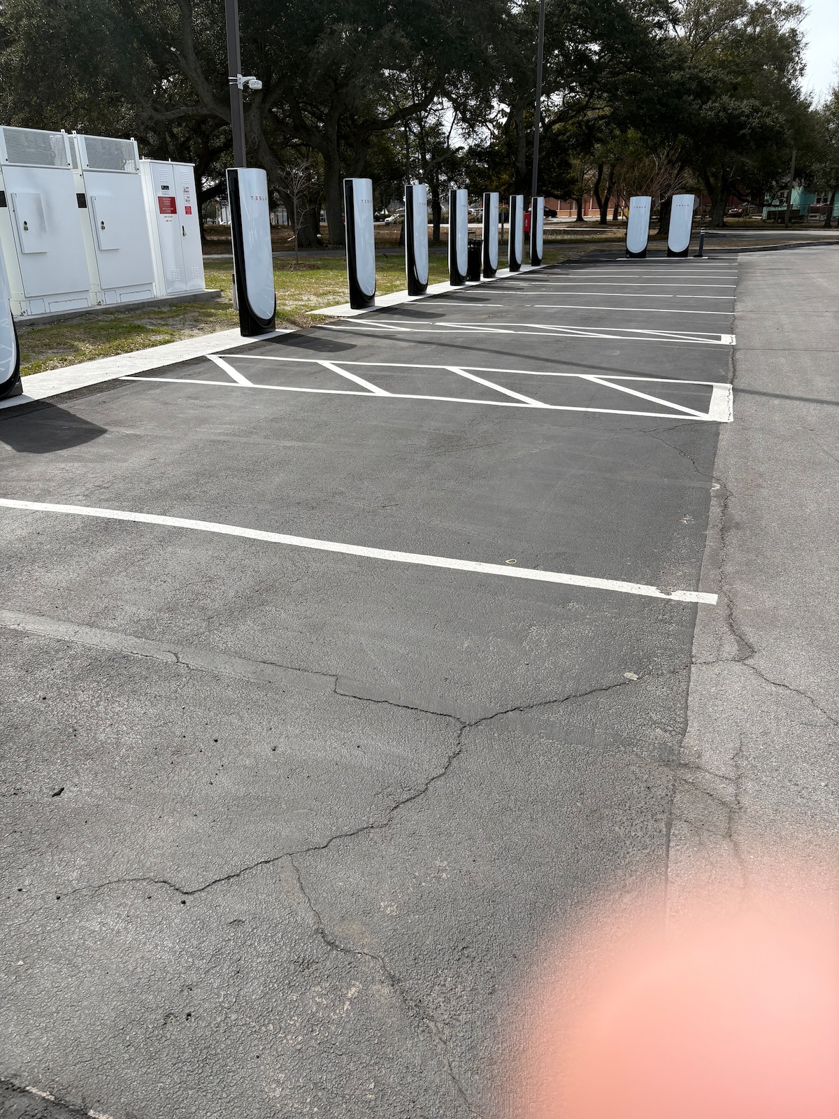 A parking lot features multiple electric car charging stations lined along one side. The blacktop surface shows clear white parking lines. The area is well-maintained, surrounded by green grass and trees, ensuring convenience for electric vehicles.