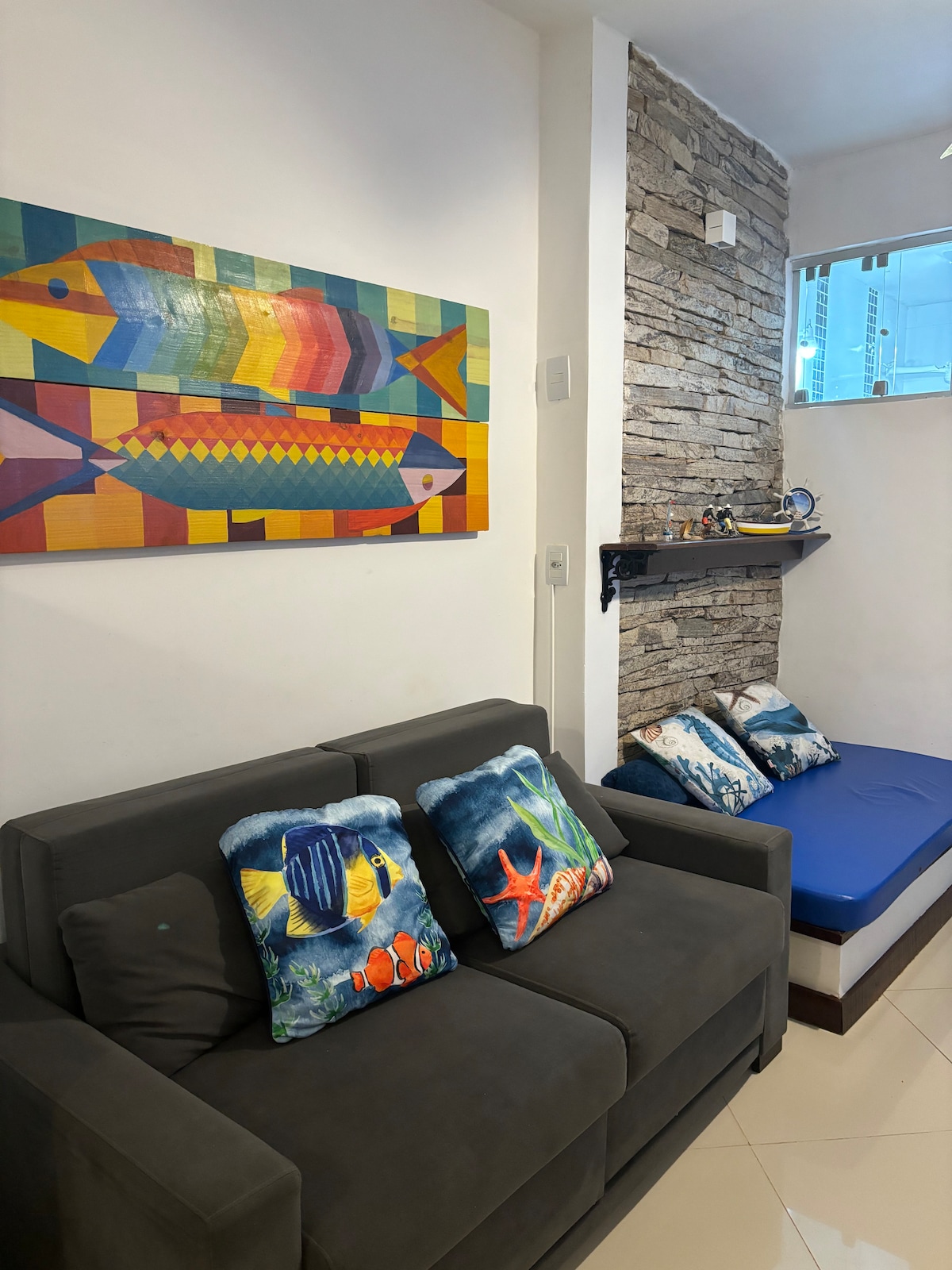 A cozy seating area is highlighted by a dark grey sofa adorned with colorful fish-themed cushions. A vibrant fish painting is displayed above, while a stone feature wall adds texture. A small nook with light blue cushions is positioned nearby, enhancing the inviting atmosphere.